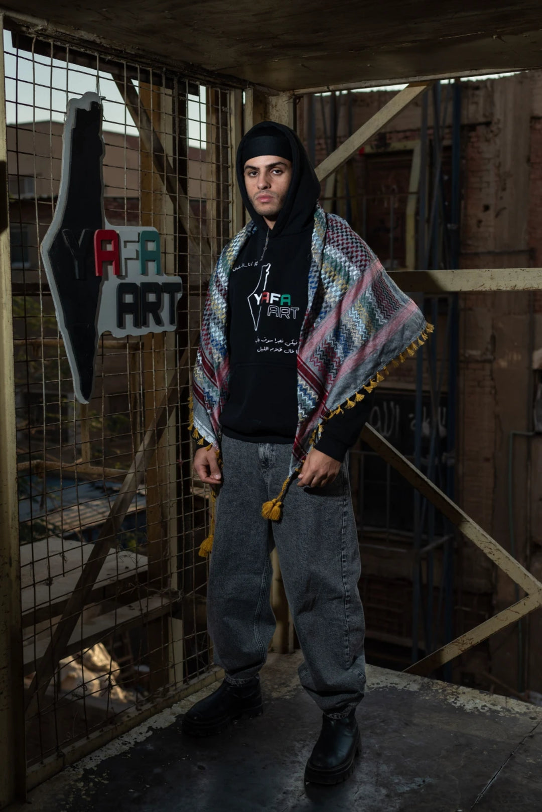 Person wearing a colorful patterned shawl over a black hoodie with text, standing in an industrial setting.